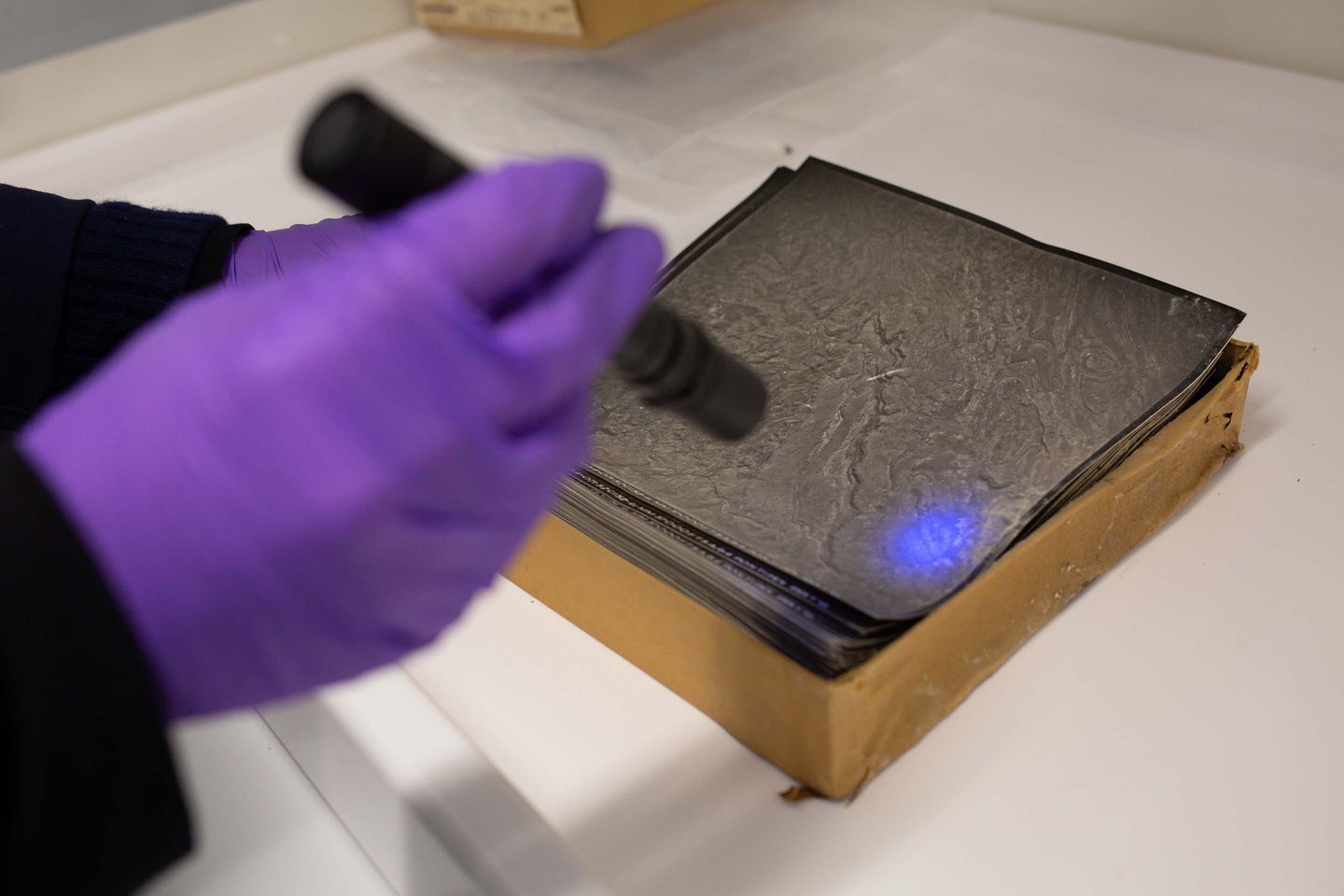 Two purple gloved hands reach into the frame from the left. The hand on the right is holding a slim black torch which shines a spot of blue light onto a black and white aerial photo. The photo is one of a stack in a cardboard box.
