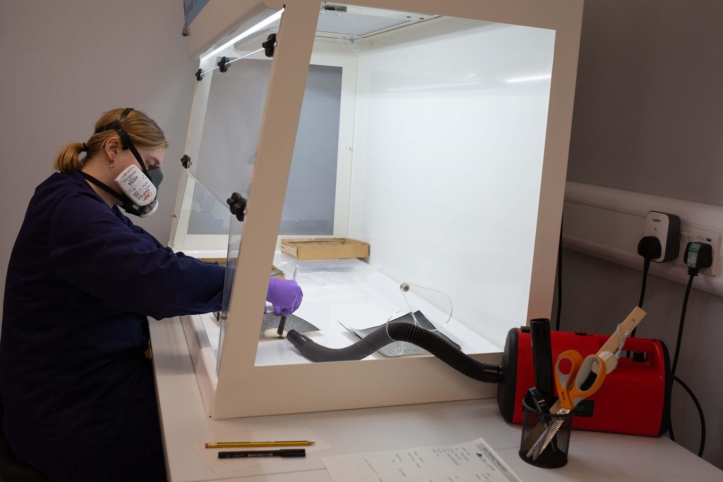 A figure in a blue lab coat wearing purple gloves and a black and white mask reaches into a fume cabinet with Perspex sides and a white frame to brush a photographic print. A red vacuum cleaner sits nearby with its hose feeding into the cabinet.