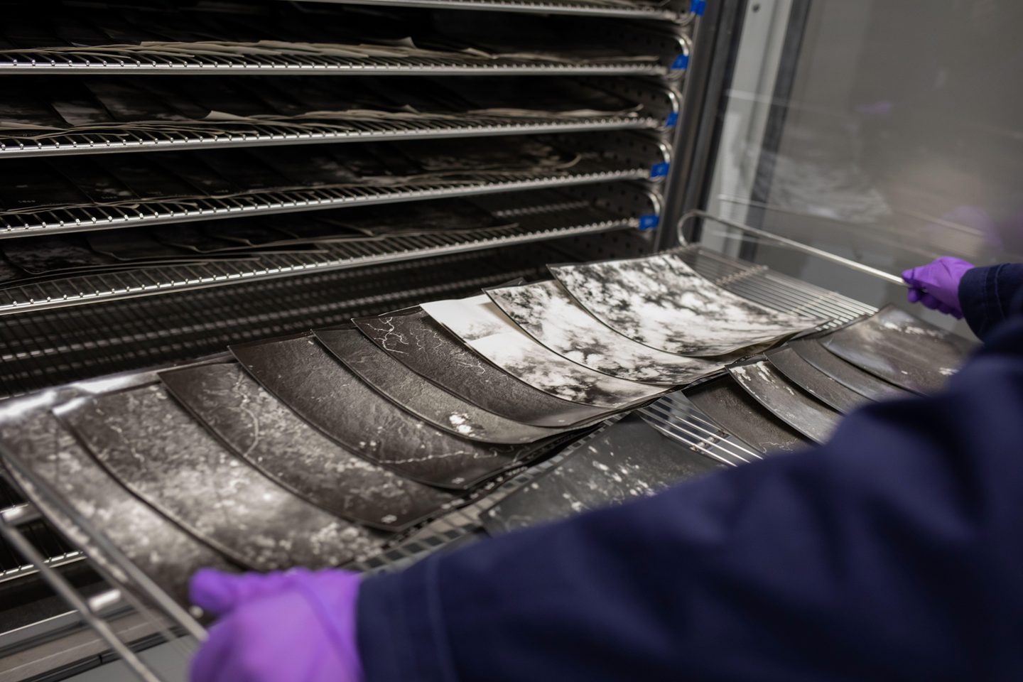 A person’s arms reach into the frame from the right side. They are wearing purple gloves and holding the edges of a tray, across which overlapping black and white aerial photos are laid. Behind this are other trays stacked on top of each other.