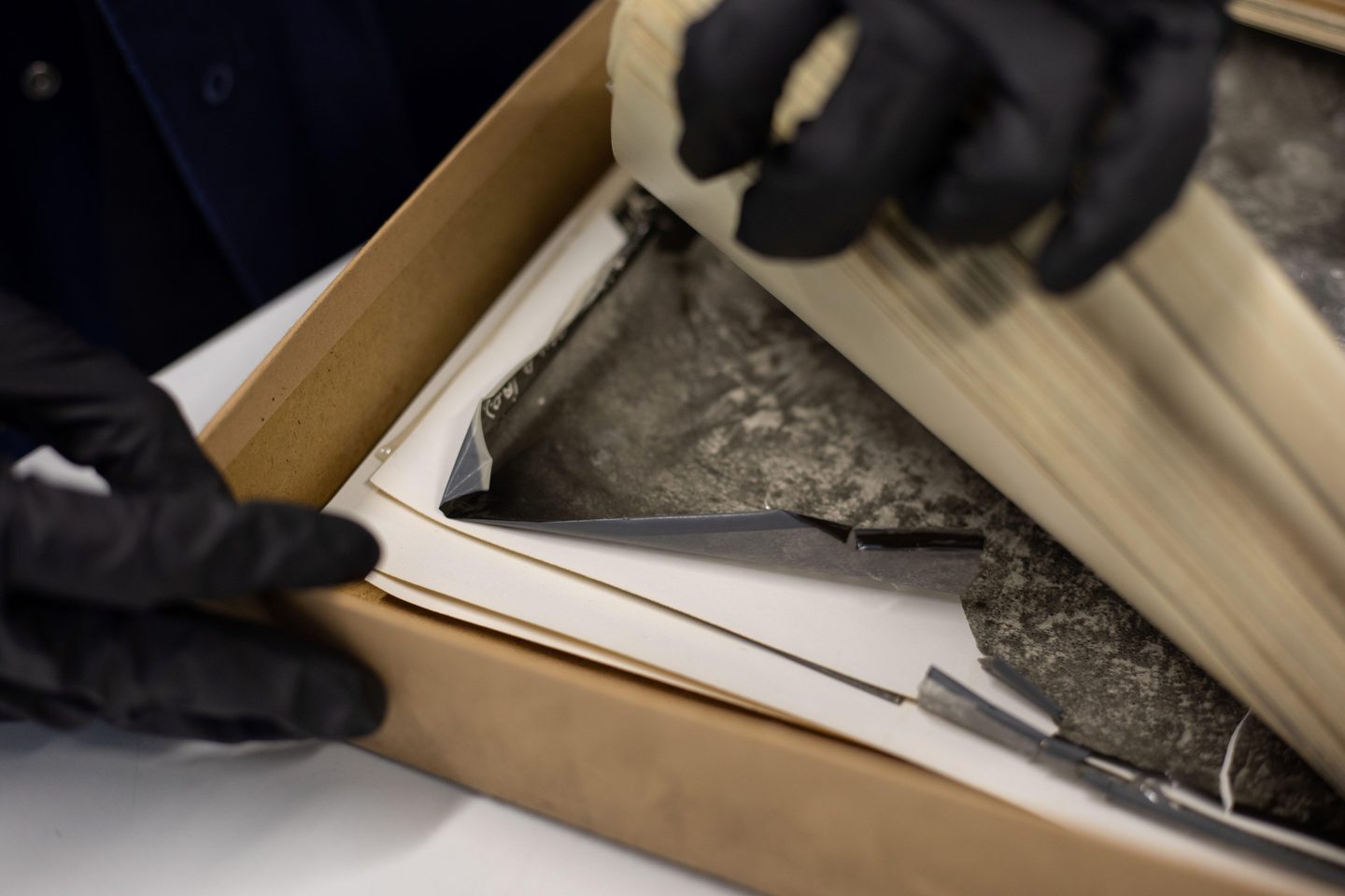 Two hands wearing black gloves enter the image from the top and the left of the frame. The top hand holds up a stack of black and white aerial photos to reveal a crumpled photo with a brittle look in a cardboard box below.