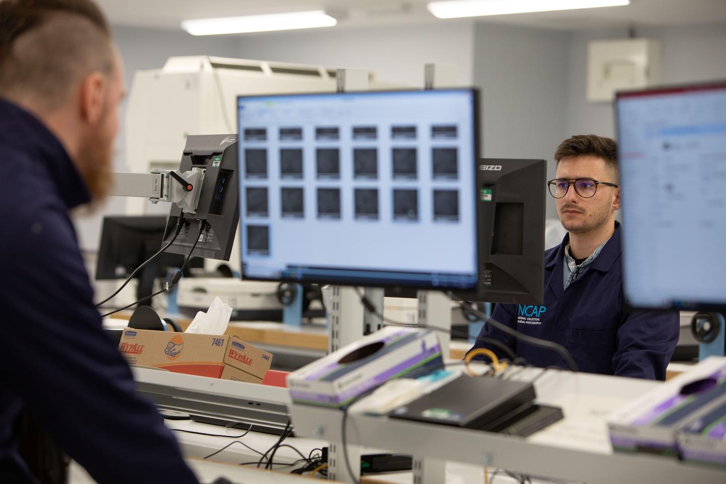 A man with short brown hair and glasses faces the camera, looking at a computer screen. Between us and the man are two out of focus screens facing us. To the left another man with cropped hair and a ginger beard faces away from us, also out of focus.