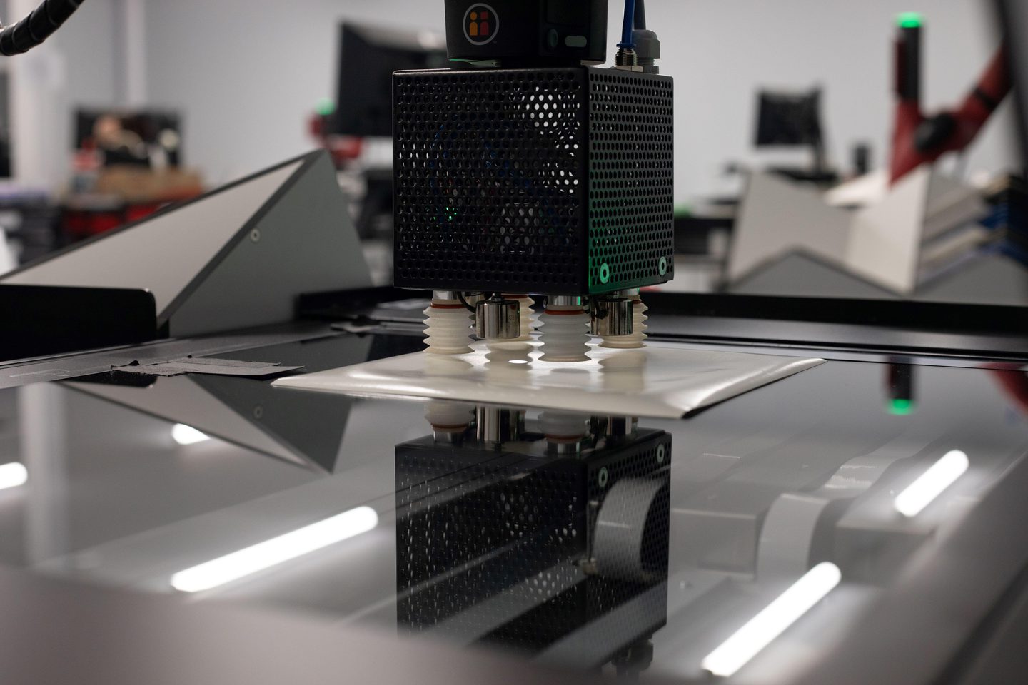 The glass surface of an image scanner is illuminated by bars of white light from below. A photographic print rests face down on the glass. A black cube with 4 white nozzles hovers over print, about to pick it up.