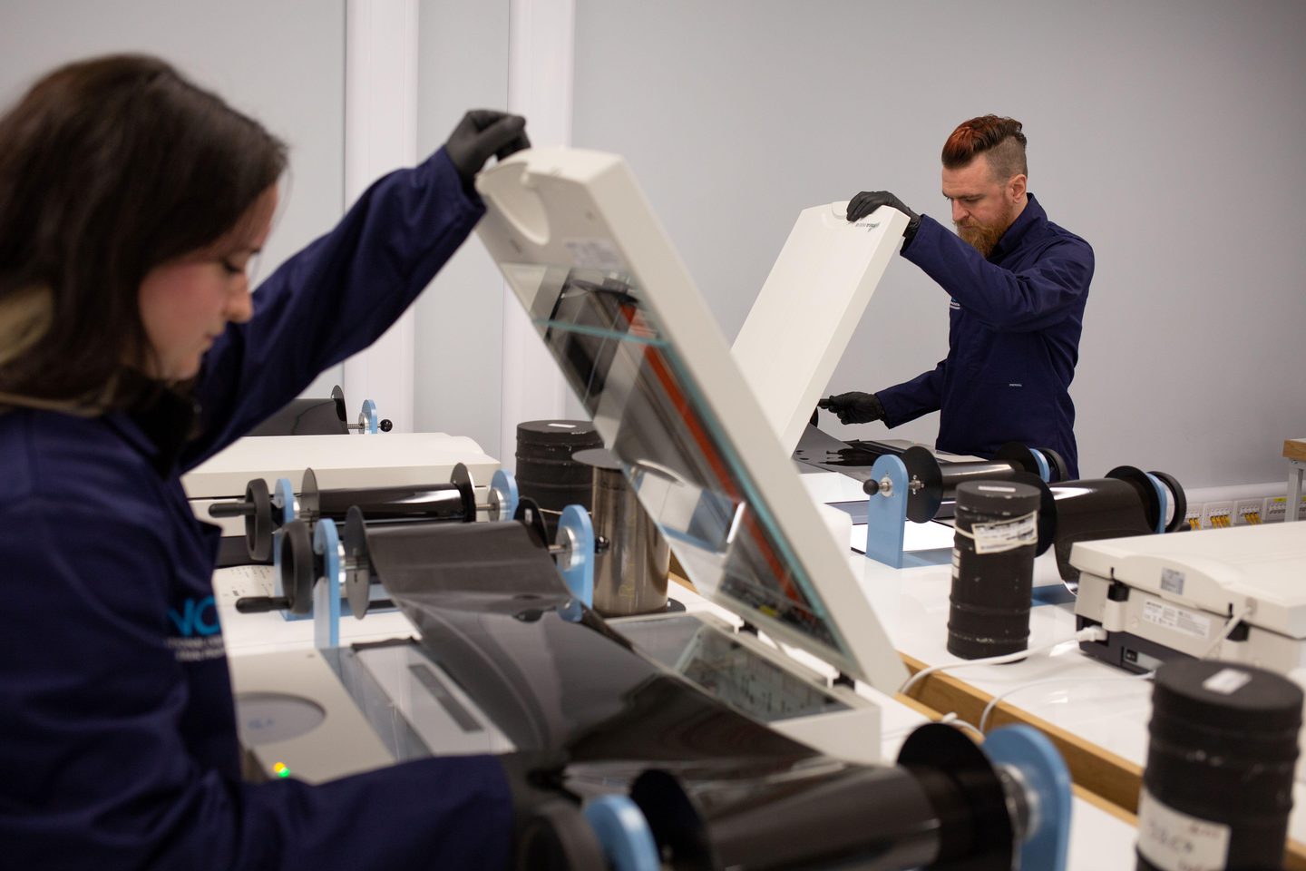 Two figures each hold open an image scanner on a white desk. They wear blue lab coats and black gloves. Each looks down and adjusts a roll of film that is being spooled across the scanner. Rolls of film and film canisters surround the scanners.
