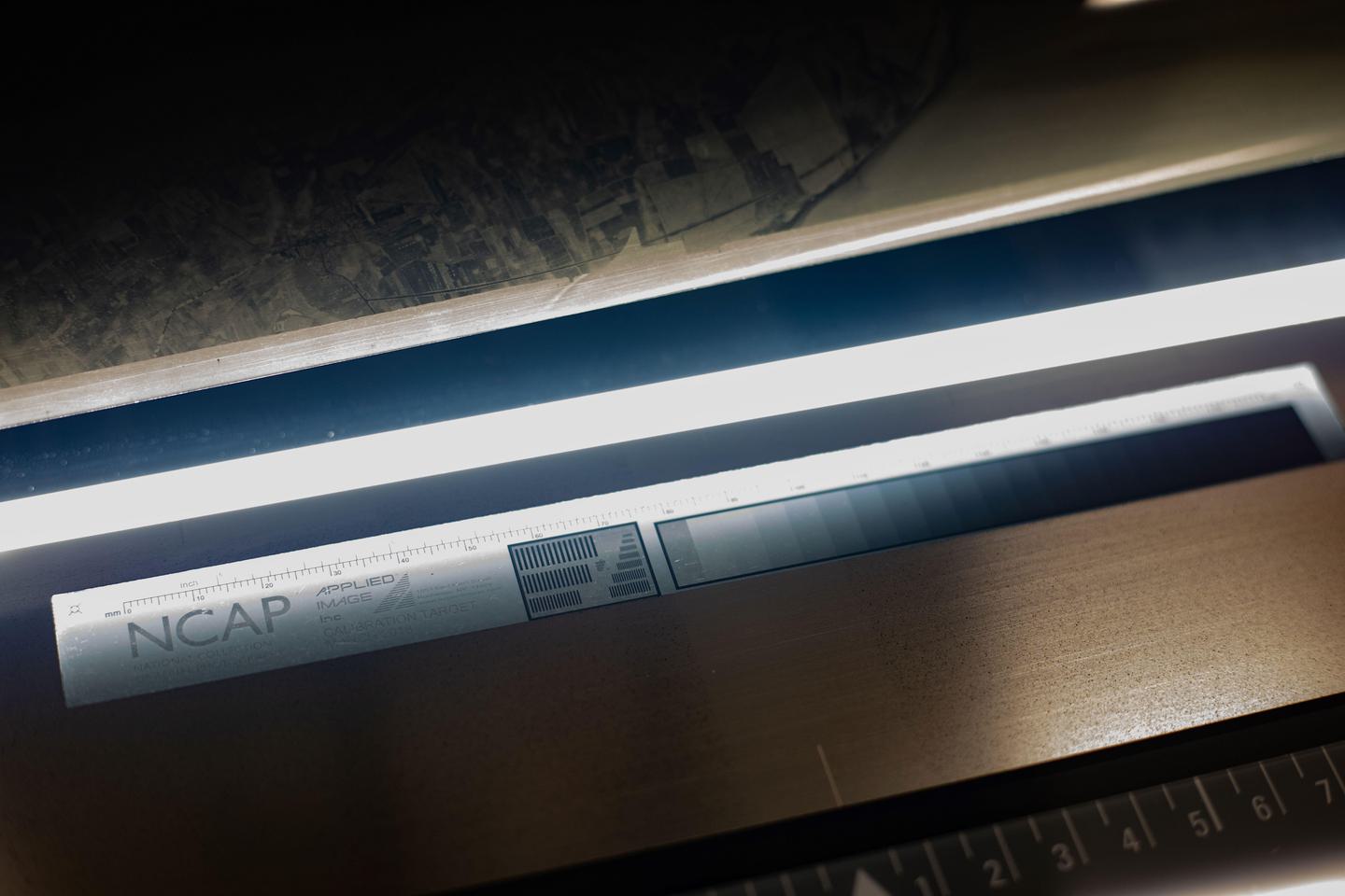 A thin strip of white light extends horizontally across the middle of the image. Above, the edge of a black and white aerial photo can be seen. Below is a label with measurement markings, a greyscale colour chart and the word ‘NCAP’.