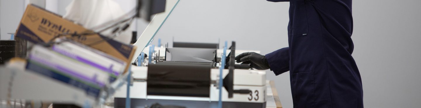 A man with dyed red hair and wearing a blue lab coat and black gloves lifts the lid of a scanner with his left hand. His right hand reaches for a black winding handle. A screen, boxes of gloves and other scanners are out of focus in the foreground.