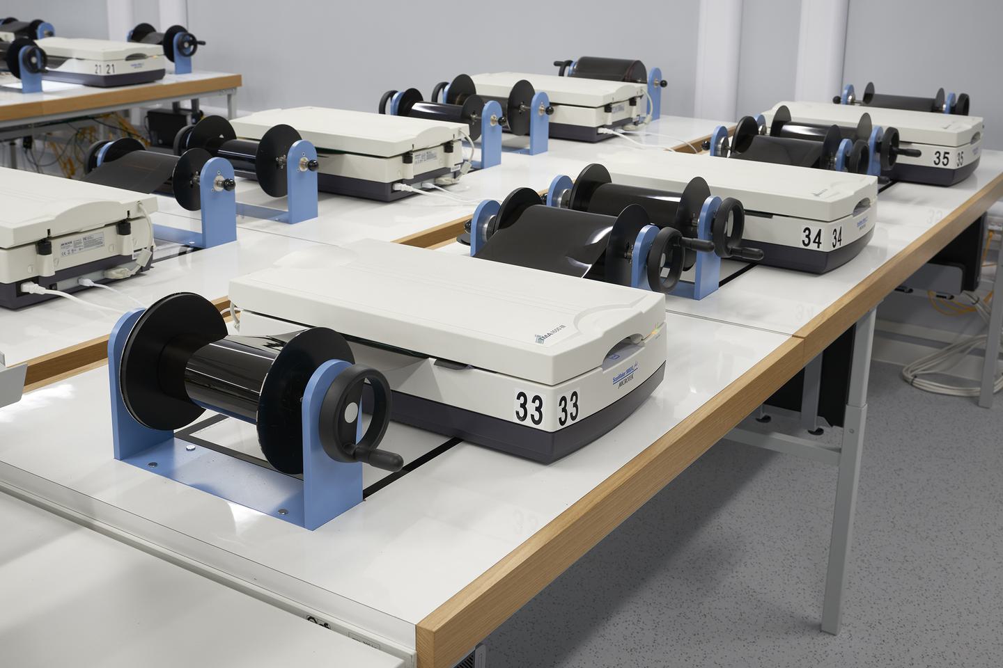 Seven white and grey image scanners spaced across lines of desks. The machines are closed. On either side of each scanner are rollers for film. The films are held on one of the rollers, fed through the scanner, then wound onto the second roller.
