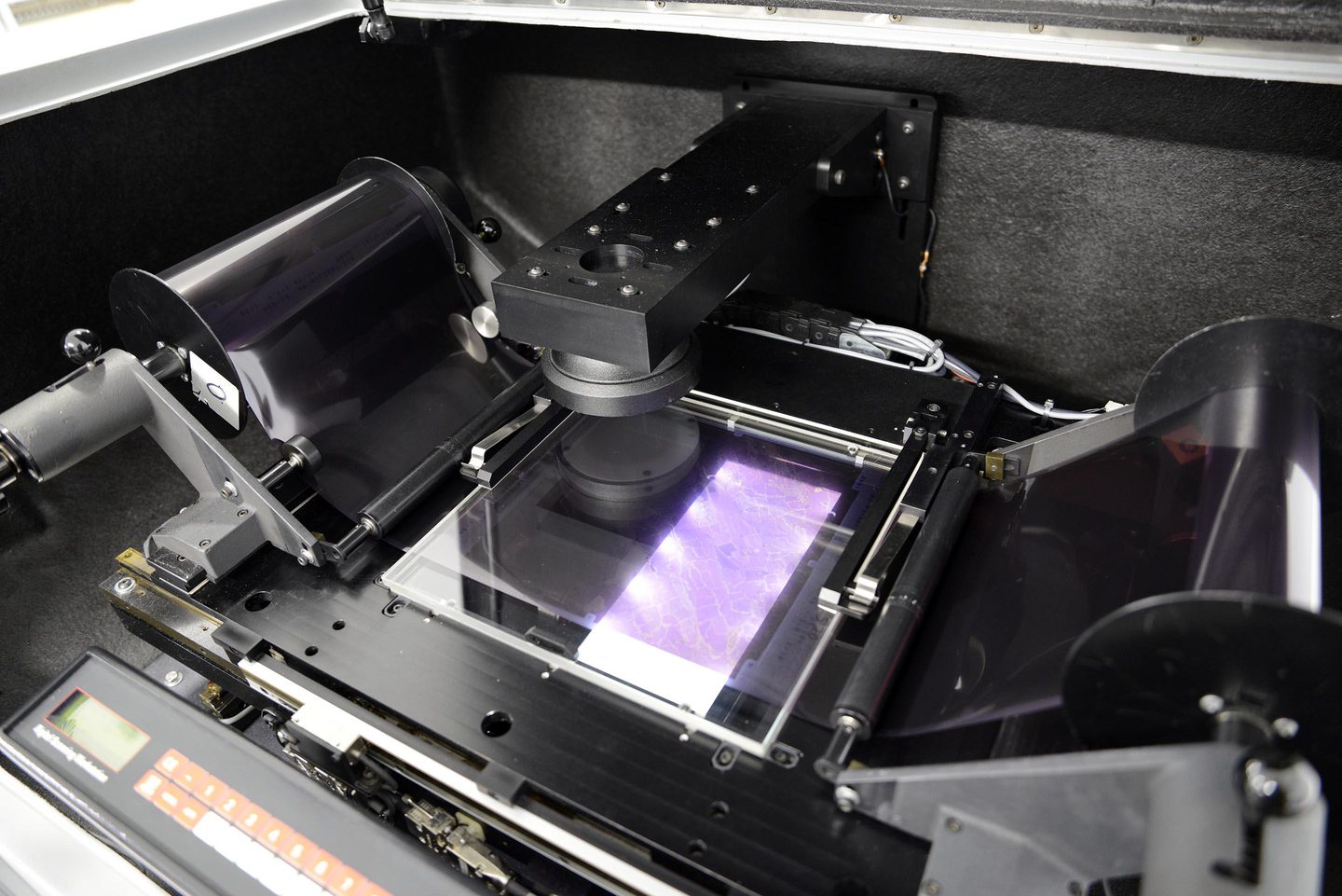 A roll of film spools across a machine, connected to rollers on each side. The central section runs over a brightly lit, glass scanning bed. On the roll the film looks dark, but the light from the scanning bed gives the film a lavender purple glow.