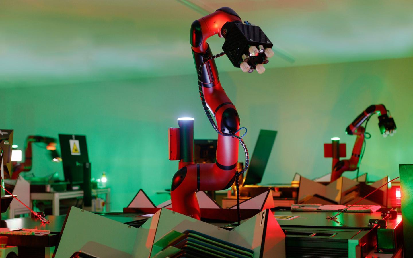A red robotic arm ending in a black cube stands vertically in the centre of the image. The arm sits between two black scanners. Other arms and scanners are visible in the background. The room is suffused with a green light.