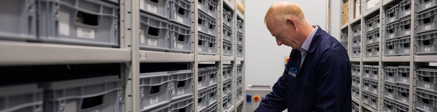 An aisle between two high racks of metal shelving. The shelving is packed with grey plastic trays. A man with red hair in a blue lab coat has pulled one of the trays half off the shelving. He has extracted a small, clear plastic box from the tray.