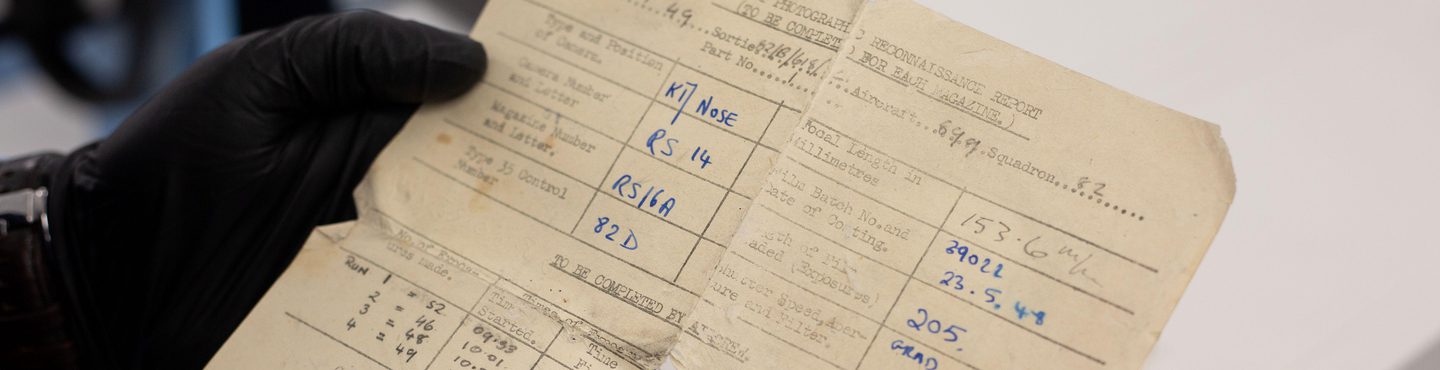 A close-up image of a piece of cream-coloured paper held by black gloved hands. It is torn on the centre fold lines and has creased and bent edges. It is printed with a form in black ink that has been completed in blue and grey handwritten text.