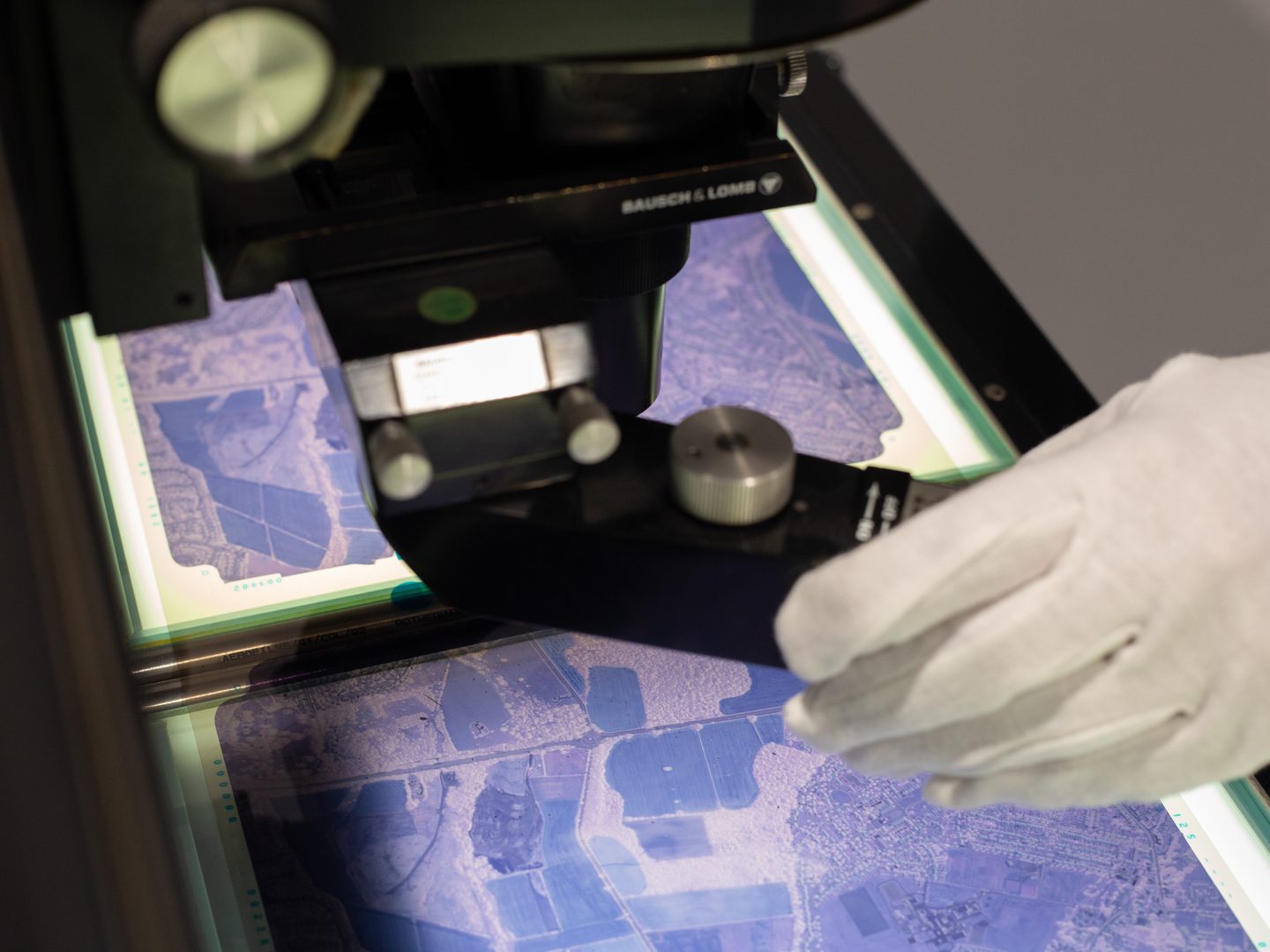 A purple-tinted aerial photographic film on a lightbox. A magnifier is suspended over the film and is being operated by a white-gloved hand.