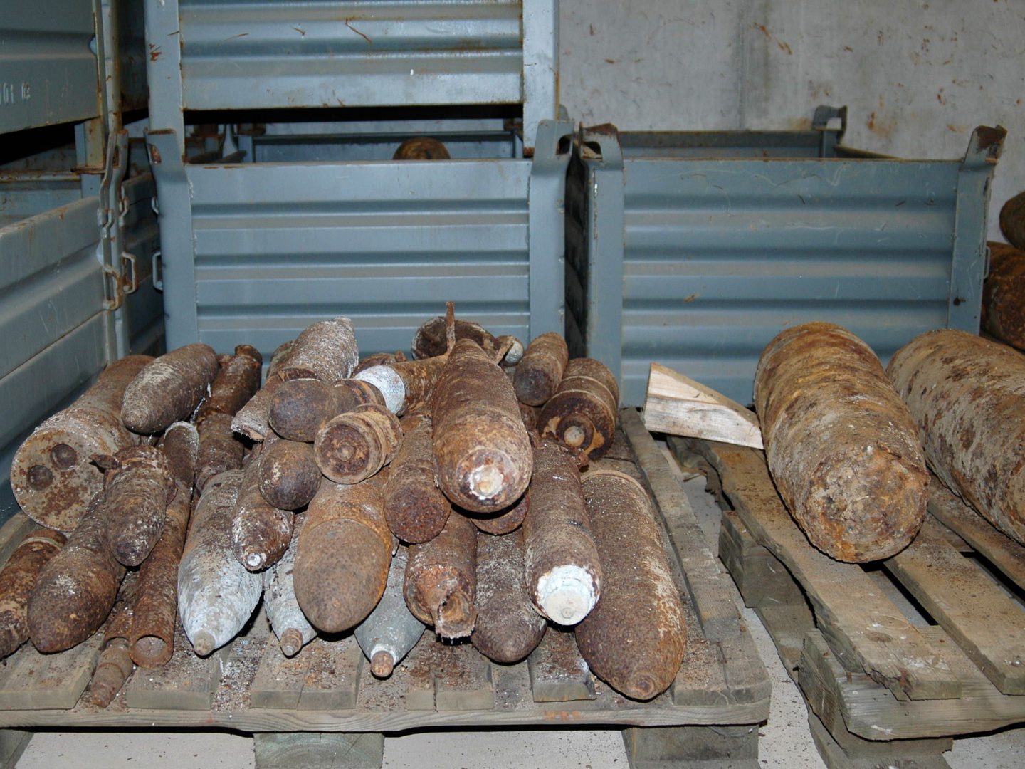 Stacks of rusted bombs on 2 wooden pallets. There are numerous small bombs on the left-hand pallet and 2 large bombs on the right-hand pallet. Grey metal containers are visible behind and to the left.