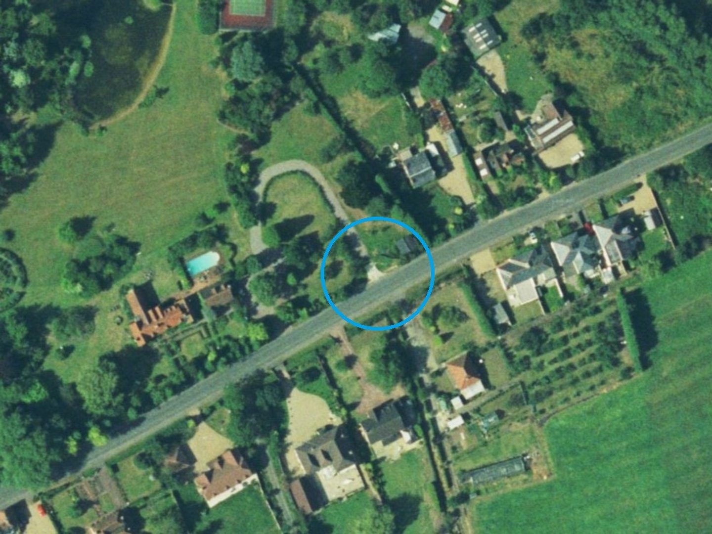 A colour aerial photo of a road flanked by houses with large gardens. It runs diagonally across the image from lower left to upper right and is surrounded by green fields and trees. A digitally drawn blue circle highlights the open end of a driveway.