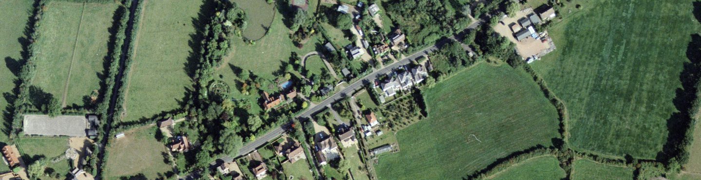 A colour aerial photo of a road flanked by houses with large gardens. It runs diagonally across the image from lower left to upper right and is surrounded by green fields, hedges and trees. A tennis court and pond are just left of centre.