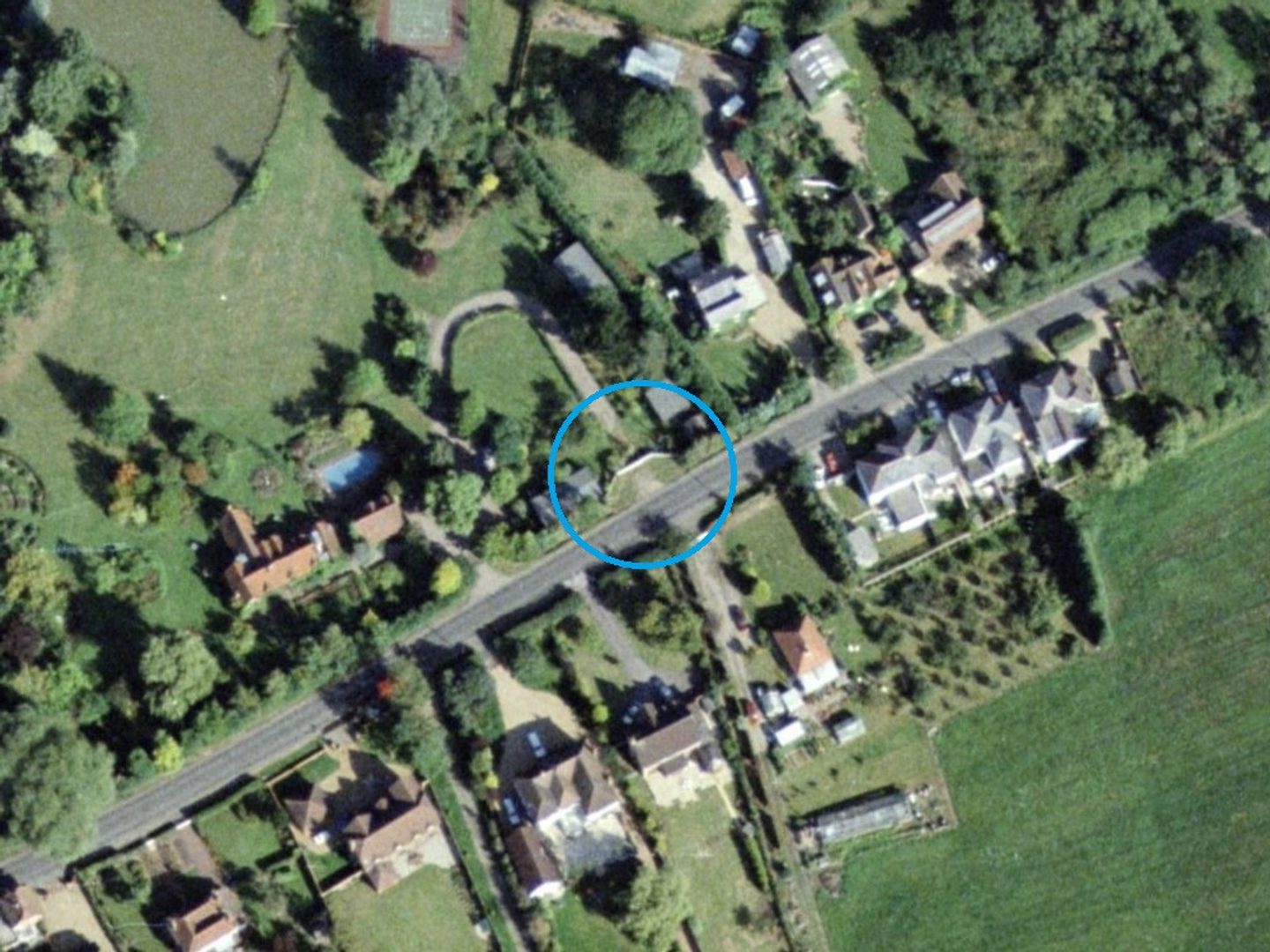 A colour aerial photo of the same road as in the previous image, running through green fields and trees from lower left to upper right. A digitally drawn blue circle highlights the same driveway as before, now blocked with a fence.