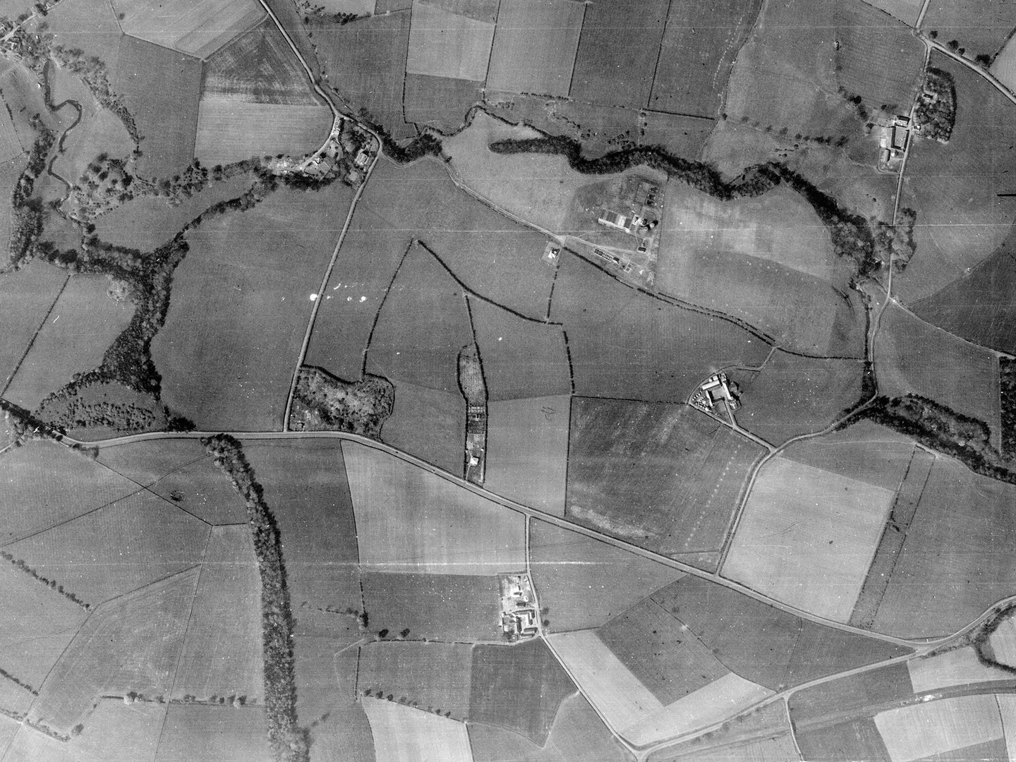 A black and white aerial photo of fields crossed by roads. The largest road runs horizontally across the middle of the image. A straight line of trees runs vertically through the left of the image, and a zig-zag line of trees runs along the top.