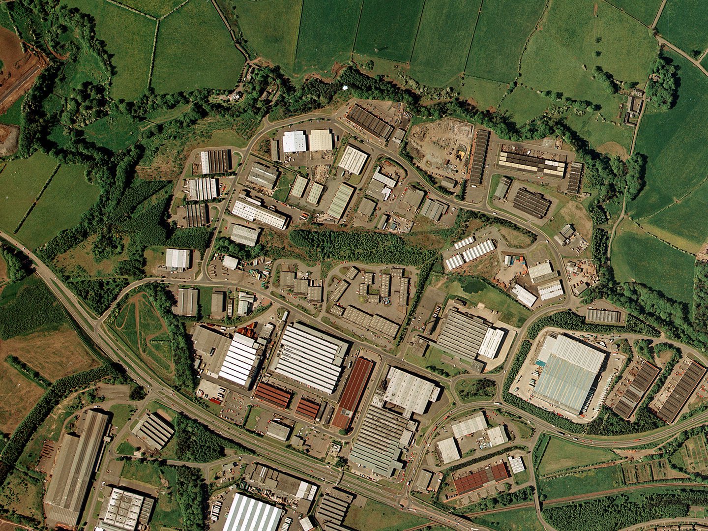 A colour aerial photo of the same scene as the image before. The lines of trees are still present but most of the fields have been built over with large industrial buildings. The scene is now crossed by a large motorway.