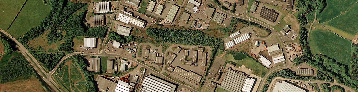 A colour aerial photo of the same scene as the image before. The lines of trees are still present but most of the fields have been built over with large industrial buildings. The scene is now crossed by a large motorway.