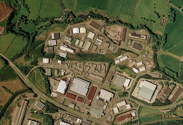 A colour aerial photo of the same scene as the image before. The lines of trees are still present but most of the fields have been built over with large industrial buildings. The scene is now crossed by a large motorway.