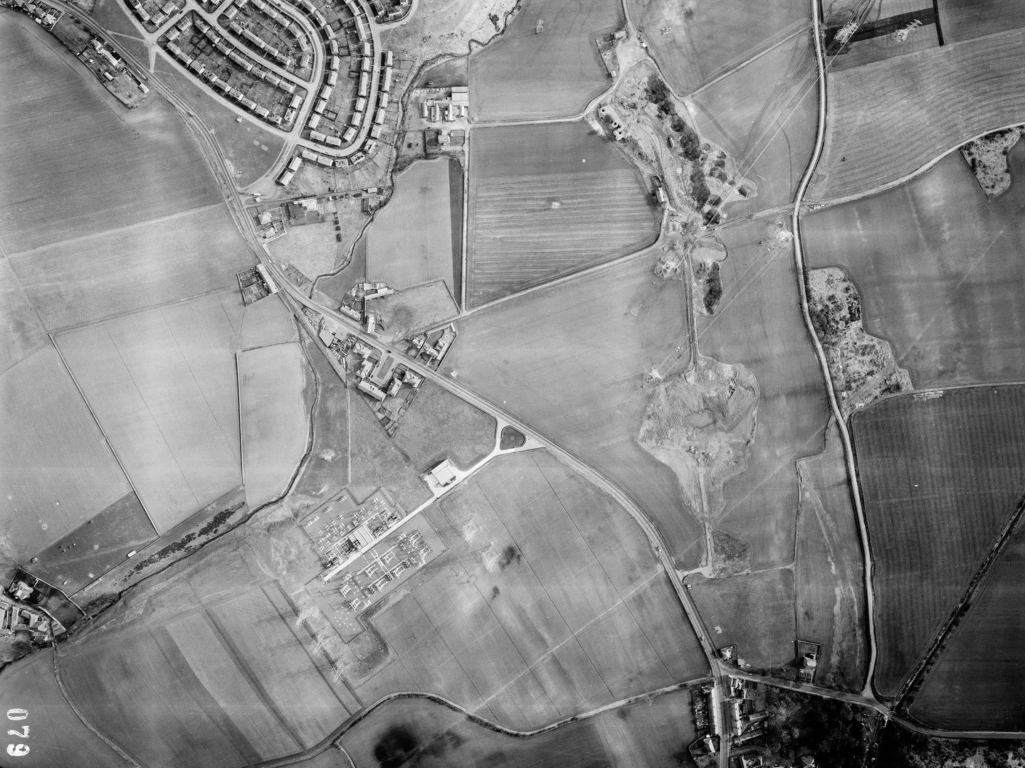 A black and white aerial photo of a landscape of fields and small woodlands crossed by a road. There are houses at the top left. In the centre and above centre are two areas of excavations. There are 3 chimneys next to the top area of excavations.