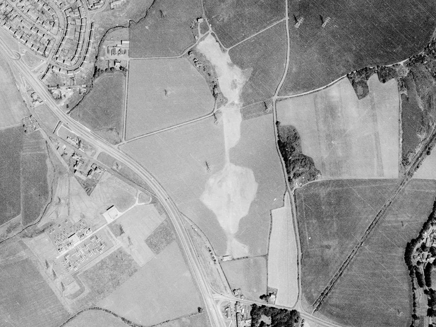 A black and white aerial photo of the same landscape of fields and small woodlands as above. There is a large bare patch in the middle of the fields where the previously visible excavations have been filled in. The road has been widened.
