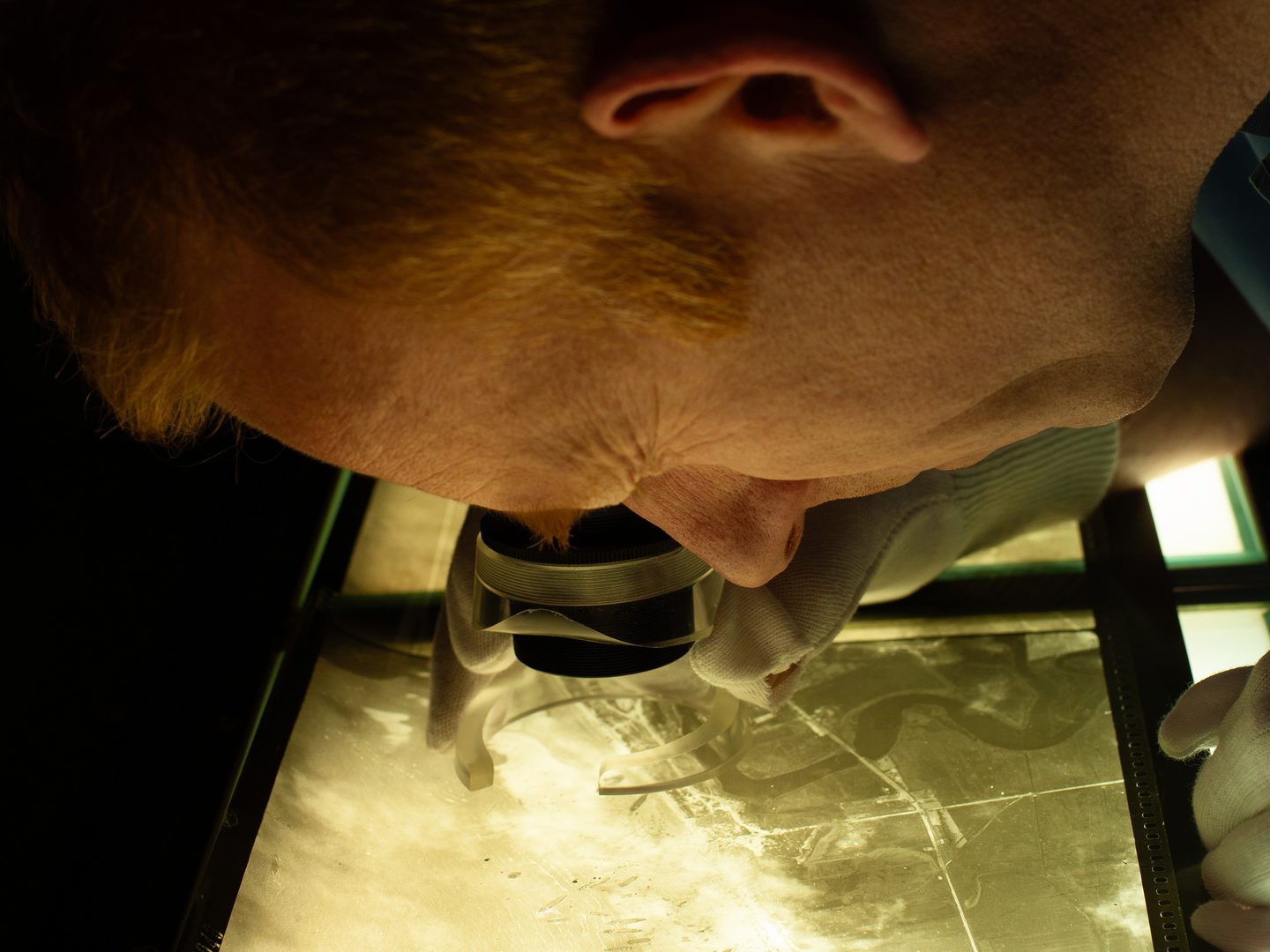 A view from the side of a man leaning over a roll of aerial photographic film, lit from below. He has one eye closed and is looking through a magnifier with the other.