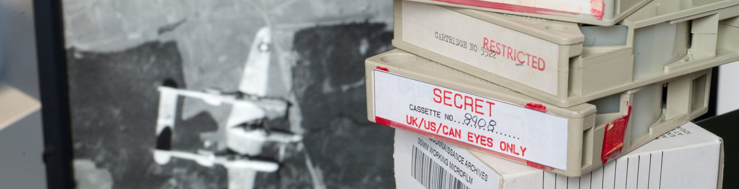 A stack of 3 microfilm cases on top of a box for storing the cases. The cases are labelled 'Secret' and 'Restricted'. In the background is an image on a screen of a Lockheed Lightning aircraft flying over a landscape.