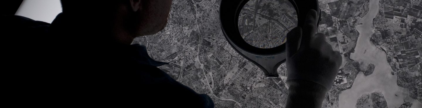 A man seen from behind leaning over a black and white aerial photo on a lightbox. He holds a magnifying lens in his white-gloved right hand.
