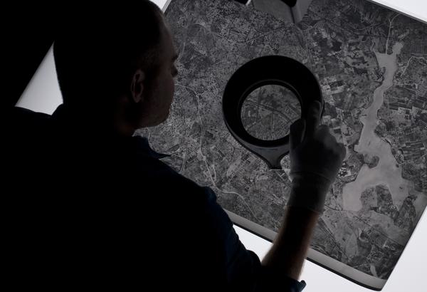 A man seen from behind leaning over a black and white aerial photo on a lightbox. He holds a magnifying lens in his white-gloved right hand.