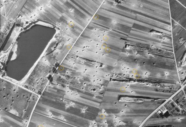A black and white aerial photo of fields. A large pond is visible centre left. The fields are pockmarked with bomb craters. Digital yellow circles have been drawn around small dots indicating where bombs have landed but not exploded.