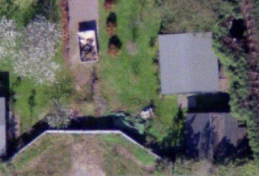 A very zoomed-in colour aerial photo of a fence built across a driveway. There are lawns either side of the driveway, and the roofs of 3 sheds are visible to the left and right.