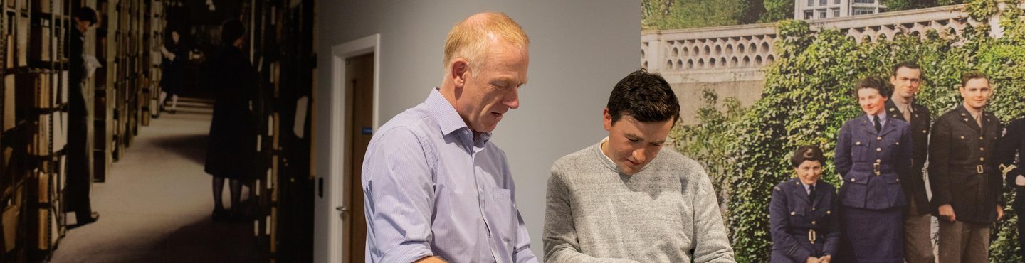 2 men examine documents they are holding. The man on the left has light red hair and wears a blue shirt. The man on the right has dark brown hair and wears a grey jumper. 2 large colourised historic photos cover the walls behind them.