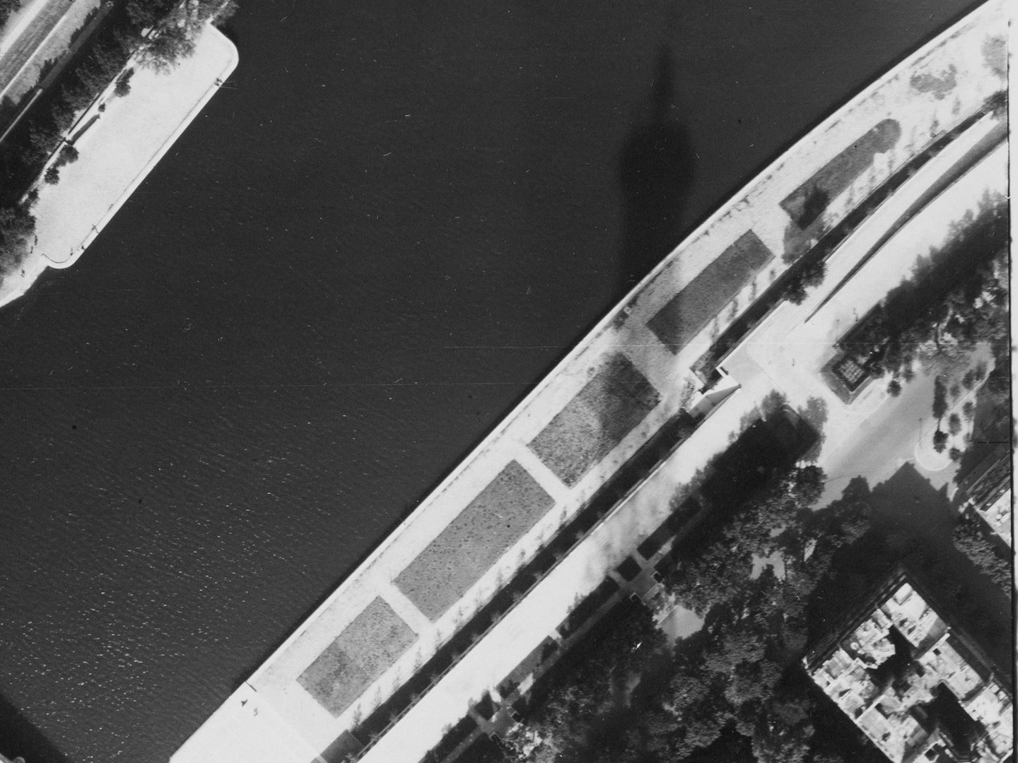 A black and white aerial photo of a river. A small area of bank is visible top left, with a larger area at bottom right. There are trees on the white banks. The sharp shadow of the Eiffel Tower stretches from top to bottom in the image’s right half.