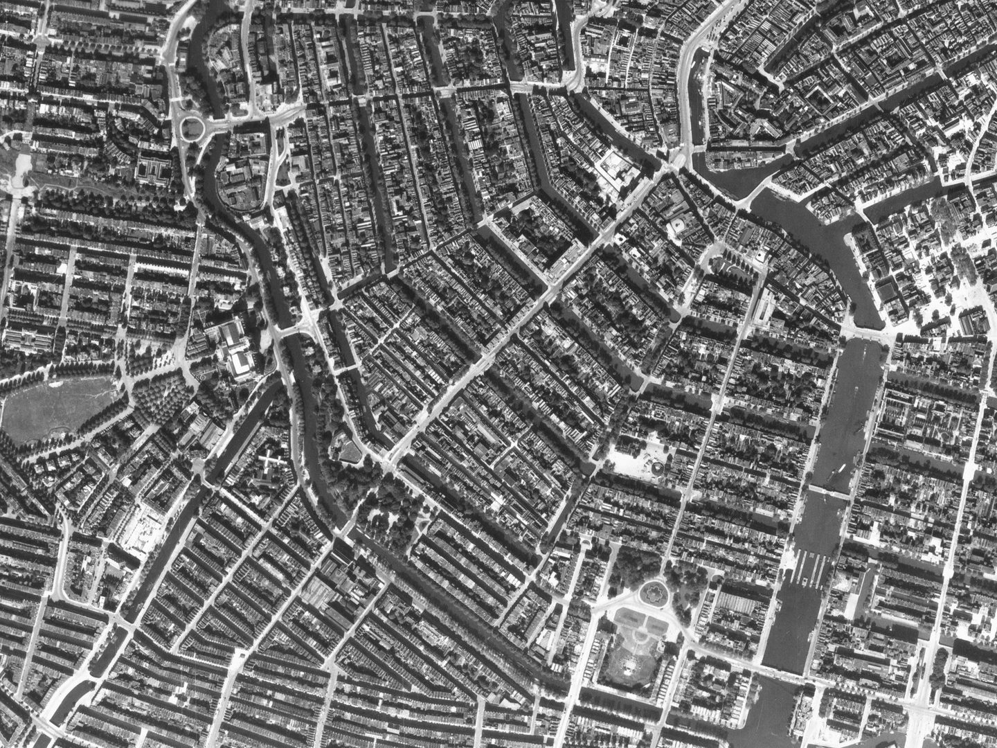 A black and white aerial photo of Amsterdam. The pattern of streets and canals is clearly visible, with a large canal running vertically through the right half of the image. The water in the canals looks black and the streets look white.