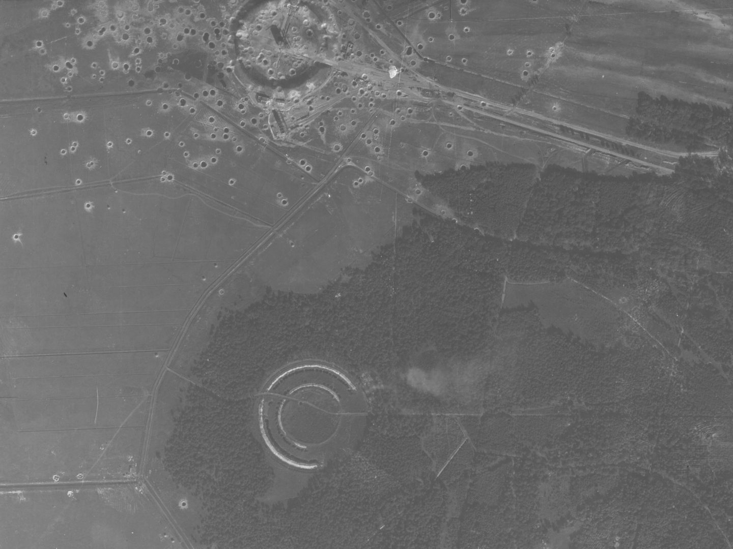 A black and white aerial photo, with the bottom and right sides taken up with a large woodland. A circular structure is visible bottom left in the woods. Another is visible at the top left surrounded by fields. It is marked with many bomb craters.