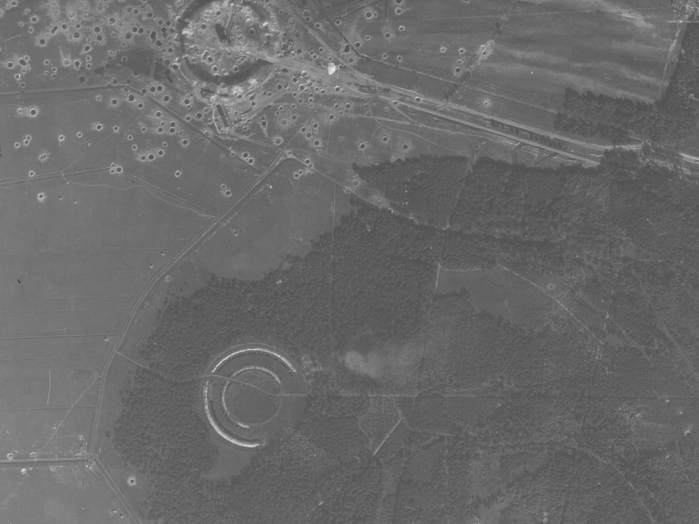 A black and white aerial photo, with the bottom and right sides taken up with a large woodland. A circular structure is visible bottom left in the woods. Another is visible at the top left surrounded by fields. It is marked with many bomb craters.