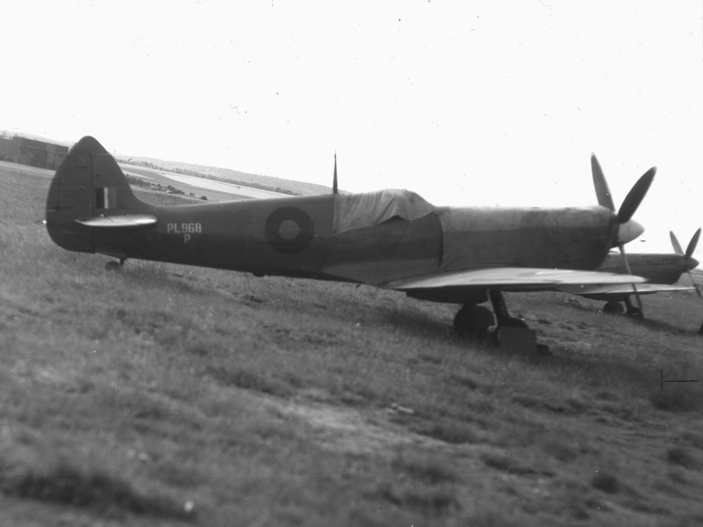 A black and white historic photo of a Spitfire plane seen from the side in a field. It is marked with the letters ‘PL968P’ and an RAF roundel. The cockpit is covered with a tarpaulin. Beyond its static propeller, a second Spitfire is just visible.