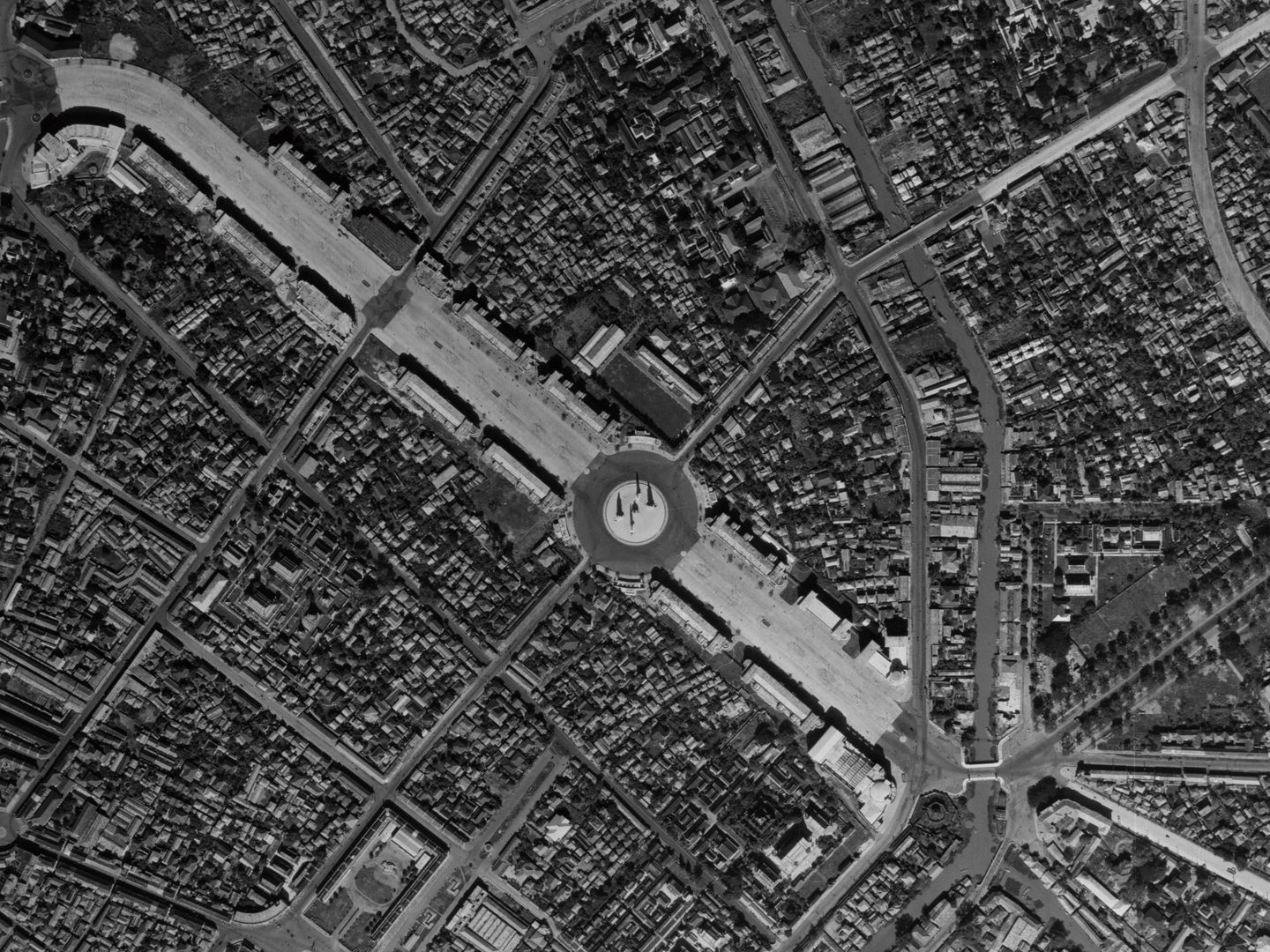 A black and white aerial photo of Bangkok. A broad boulevard runs from the top left corner to a large roundabout in the middle of the image. Buildings are densely packed either side of the boulevard. There is a smaller road and waterway to the right.