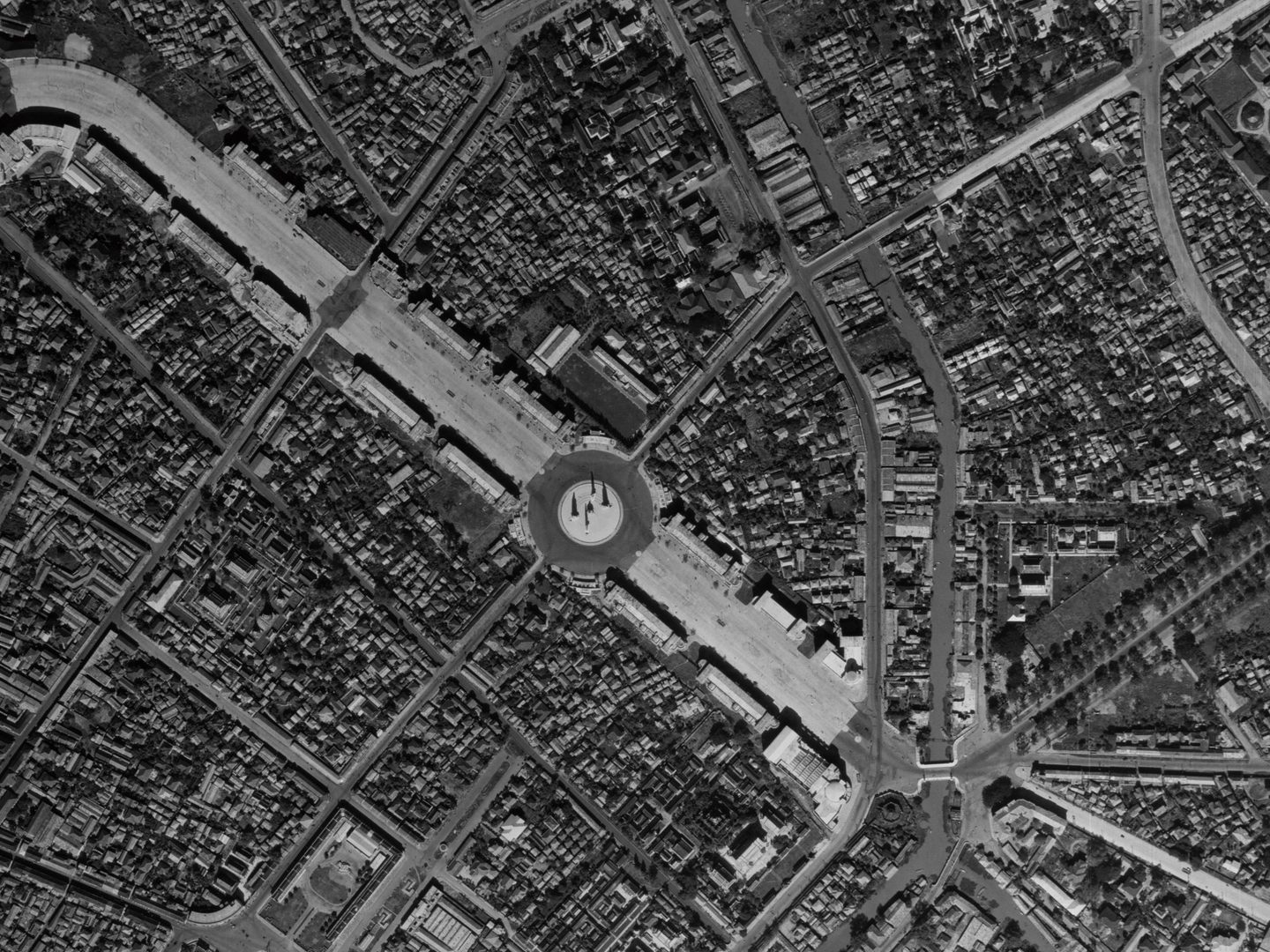 A black and white aerial photo of Bangkok. A broad boulevard runs from the top left corner to a large roundabout in the middle of the image. Buildings are densely packed either side of the boulevard. There is a smaller road and waterway to the right.