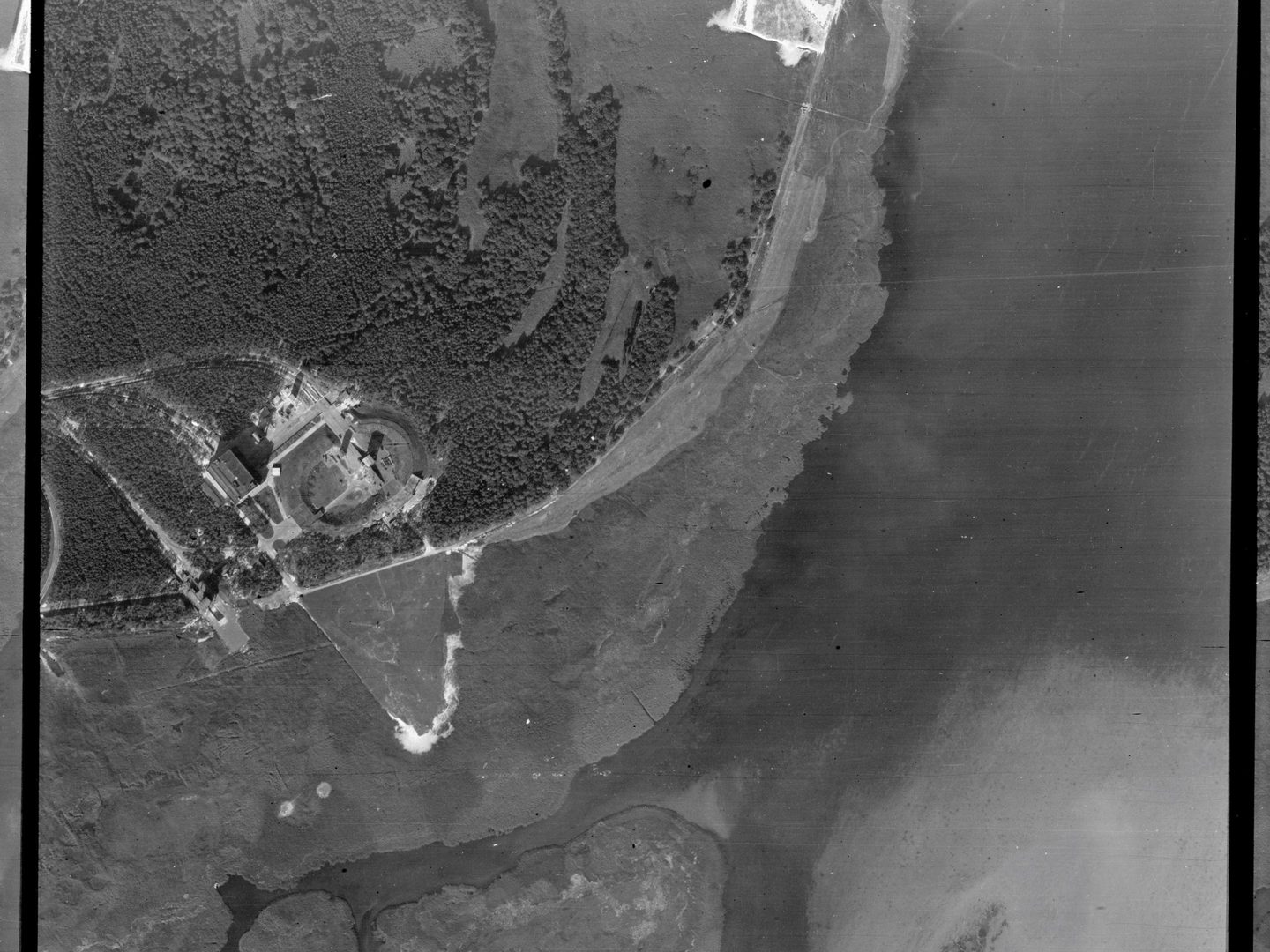 A black and white aerial photo of the same location as the image above but taken from further to the right. The woodland, buildings and oval construction are now in the top left corner. The right half and most of the bottom of the image show the sea.