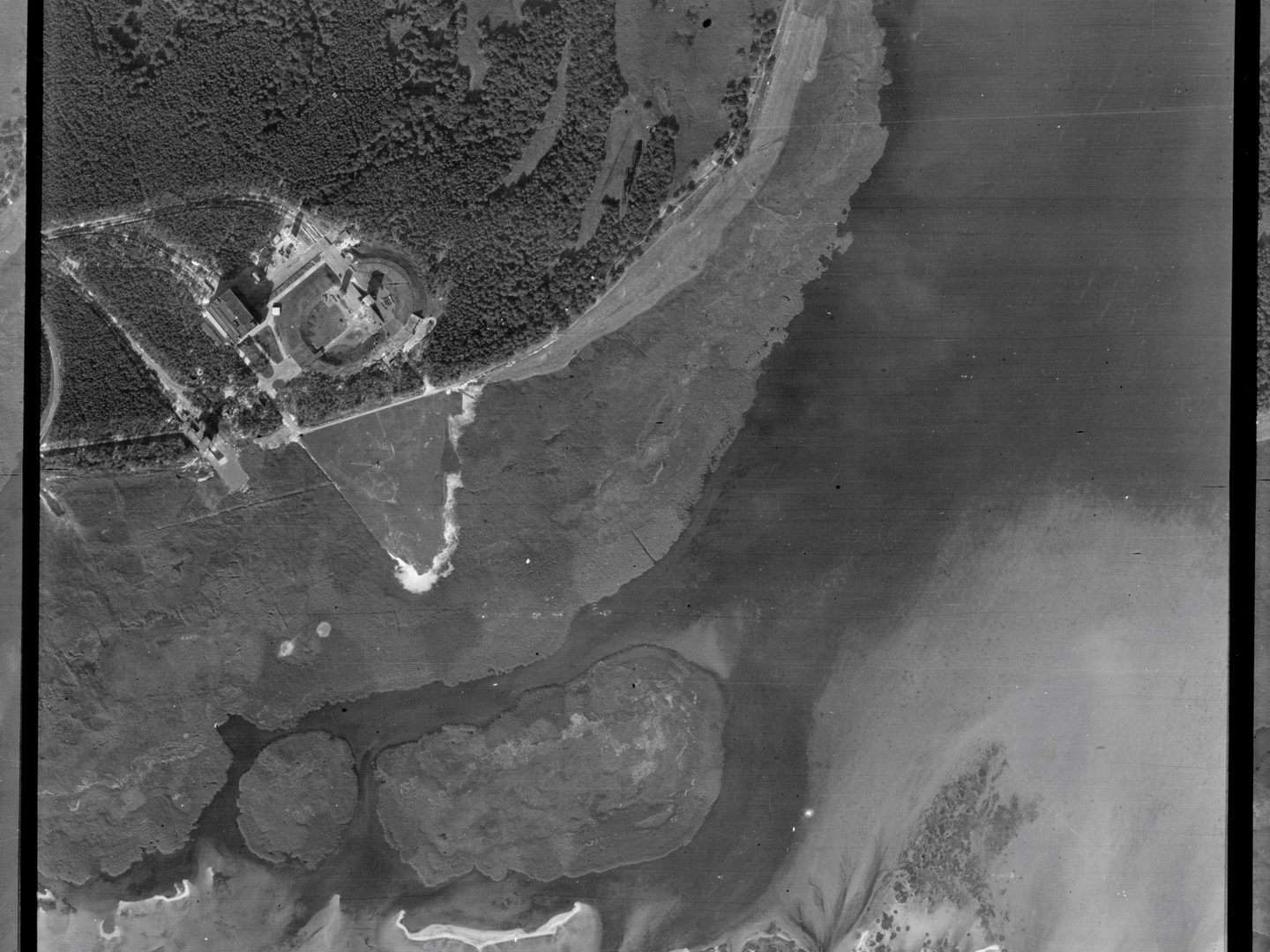 A black and white aerial photo of the same location as the image above but taken from further to the right. The woodland, buildings and oval construction are now in the top left corner. The right half and most of the bottom of the image show the sea.