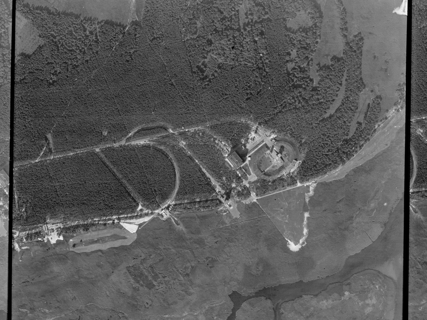 A black and white aerial photo, with a large woodland occupying the top half and a mix of open country and water in the bottom half. Several roads curve through the woodland. To the right of centre are several buildings and an oval construction.
