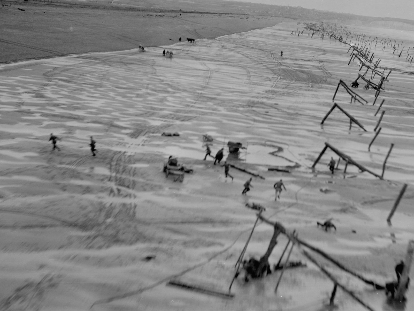 A black and white oblique aerial photo of a beach, taken from very low altitude. The beach is scattered with numerous wooden structures to make landing harder. People run for cover from the reconnaissance plane or dive to the floor in panic.