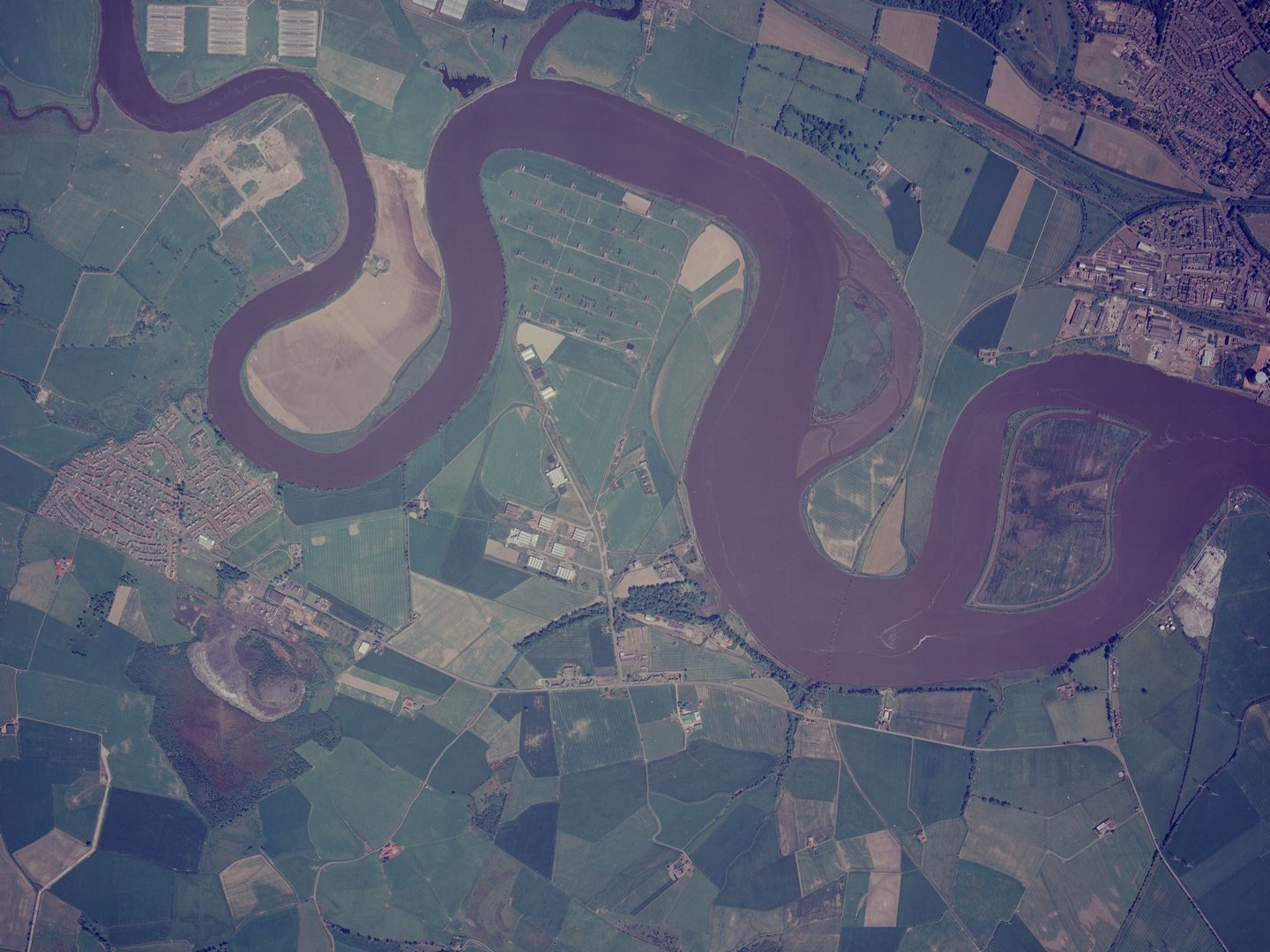 A colour aerial photo of a broad, winding river. The river is dark brown and passes between green fields. An island sits in a particularly wide part of the river. A village is visible at the centre left edge, and a town is in the top right corner.