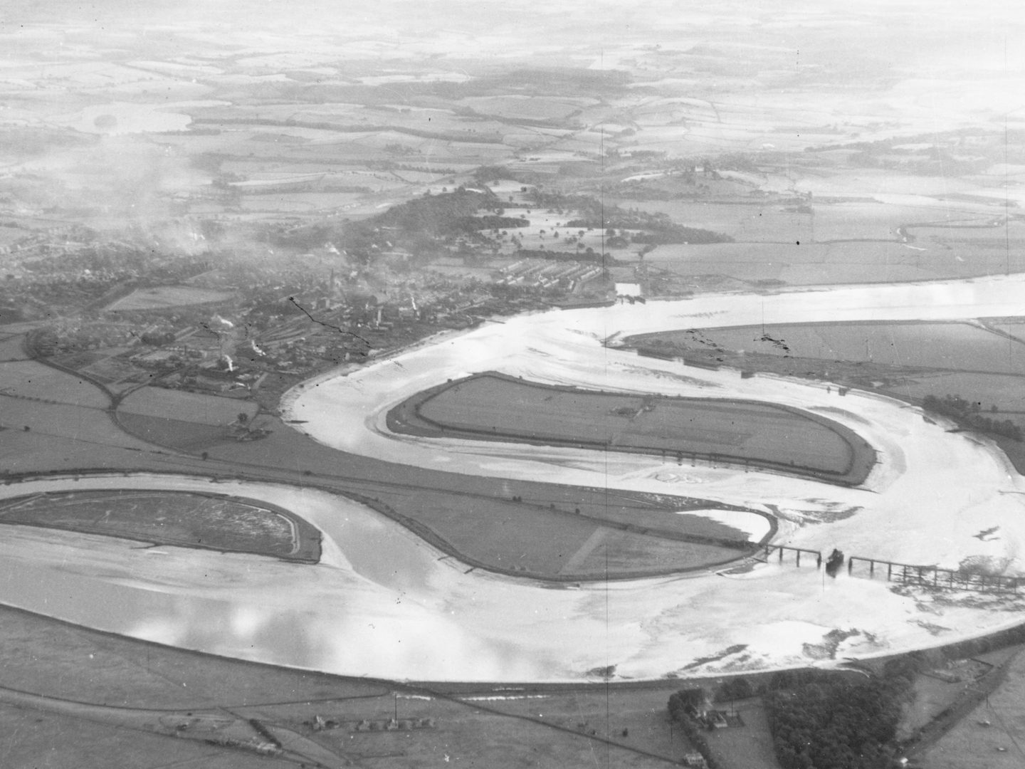 A black and white oblique aerial photo of the same river as in the above image. The same island is visible in the middle, with the town to the left. Fields stretch into the distance. A bridge is visible crossing the river in the foreground.