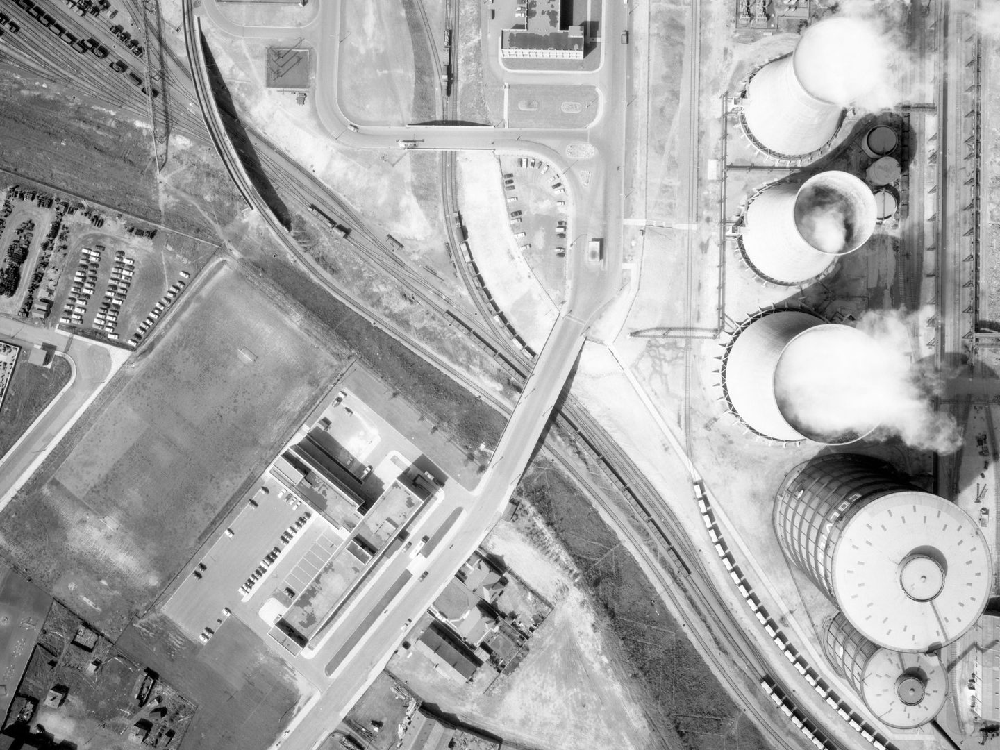 A black and white aerial photo of Ravenscraig Steel Works. A railway runs from the top left corner to bottom right. To the left are 2 carparks and some buildings. To the right are 3 cooling towers belching white steam, and a cylindrical gas holder.