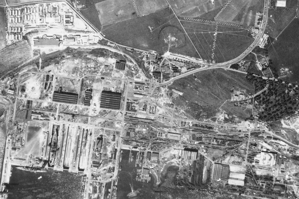 A black and white aerial photo of Rosyth docks. The image is busy and confusing. The sea is at the bottom edge with some ships in dry docks or tied up at piers. There are numerous industrial buildings and roads. At centre right is a small woodland.