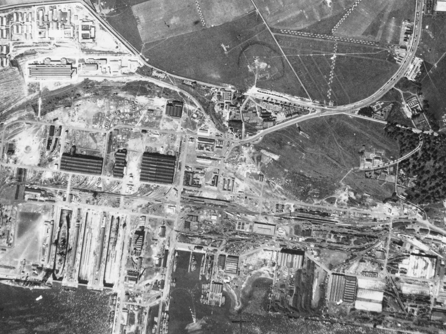 A black and white aerial photo of Rosyth docks. The image is busy and confusing. The sea is at the bottom edge with some ships in dry docks or tied up at piers. There are numerous industrial buildings and roads. At centre right is a small woodland.
