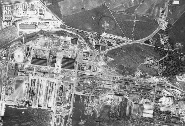 A black and white aerial photo of Rosyth docks. The image is busy and confusing. The sea is at the bottom edge with some ships in dry docks or tied up at piers. There are numerous industrial buildings and roads. At centre right is a small woodland.
