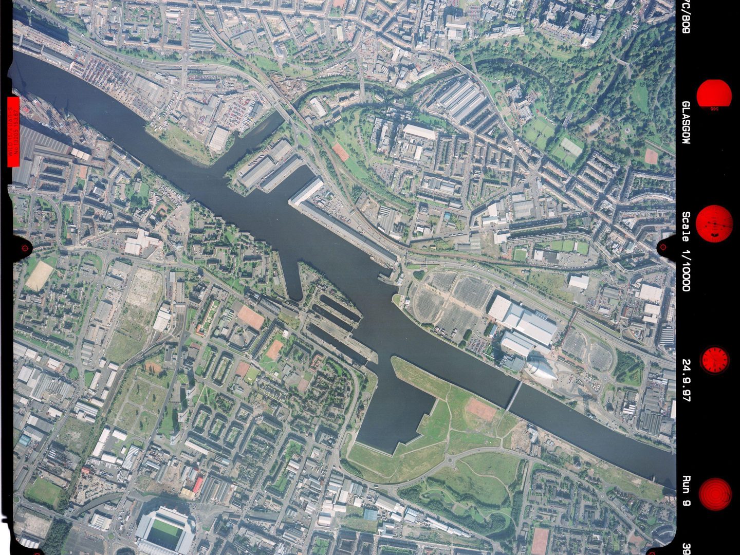 A medium-scale colour aerial photo of Glasgow. The river from the previous image now occupies the centre, running diagonally from top left to bottom right. There are industrial buildings on the riverbank, with houses and parks further away.